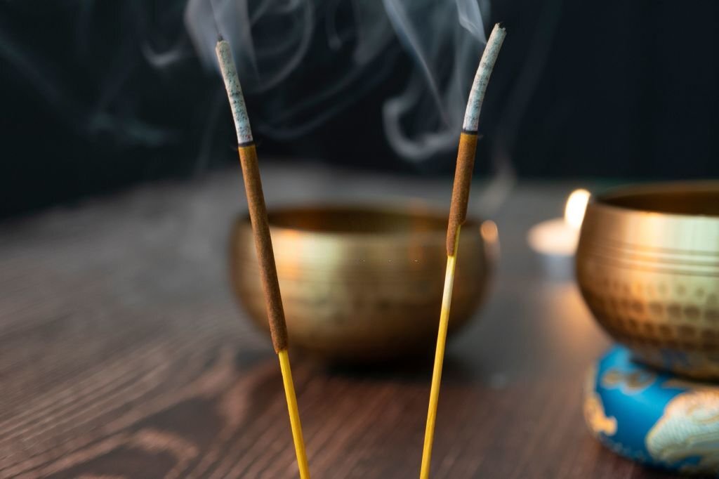 A closeup shot of two burning sticks with smoke, and a golden bowl in the background